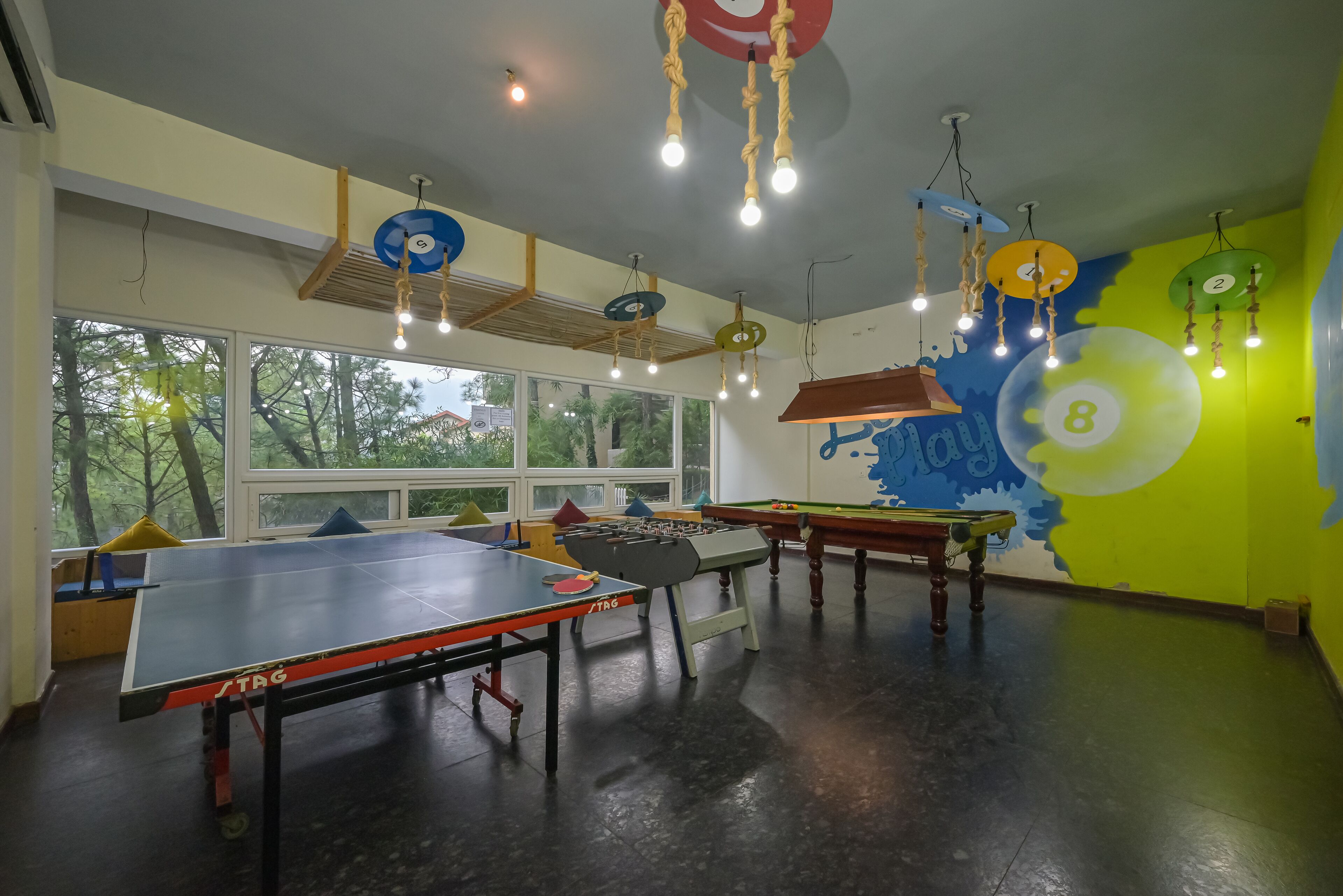Um quarto com mesa de pingue-pongue, mesa de futebol de mesa e mesa de bilhar. Há grandes janelas e um mural na parede.