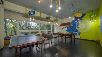 Um quarto com mesa de pingue-pongue, mesa de futebol de mesa e mesa de bilhar. Há grandes janelas e um mural na parede.