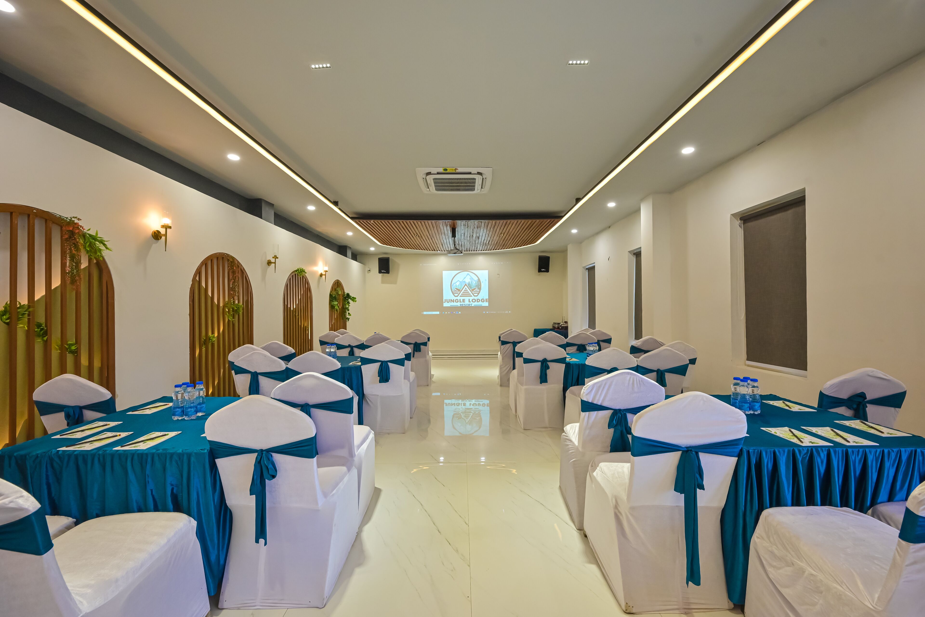 A conference room with teal and white decor, featuring tables with water bottles and napkins, and a presentation screen at the front.