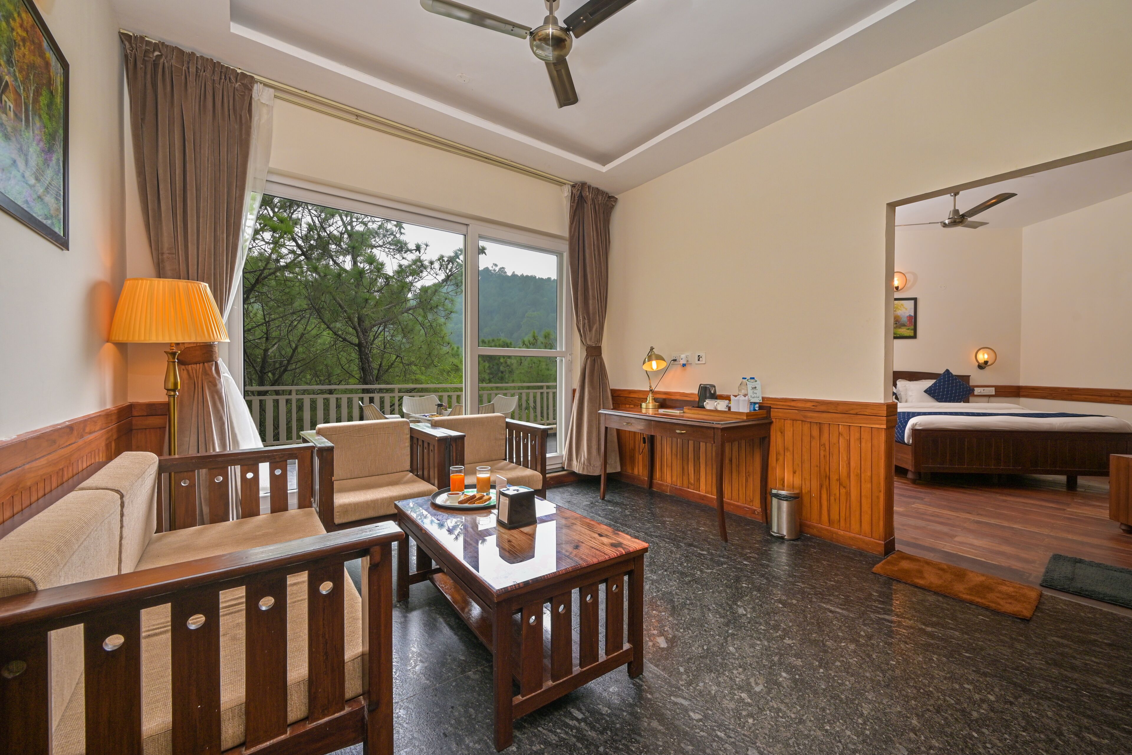 Habitación de hotel con una cama de madera, un sofá, una mesa de comedor y vistas a árboles por la ventana.