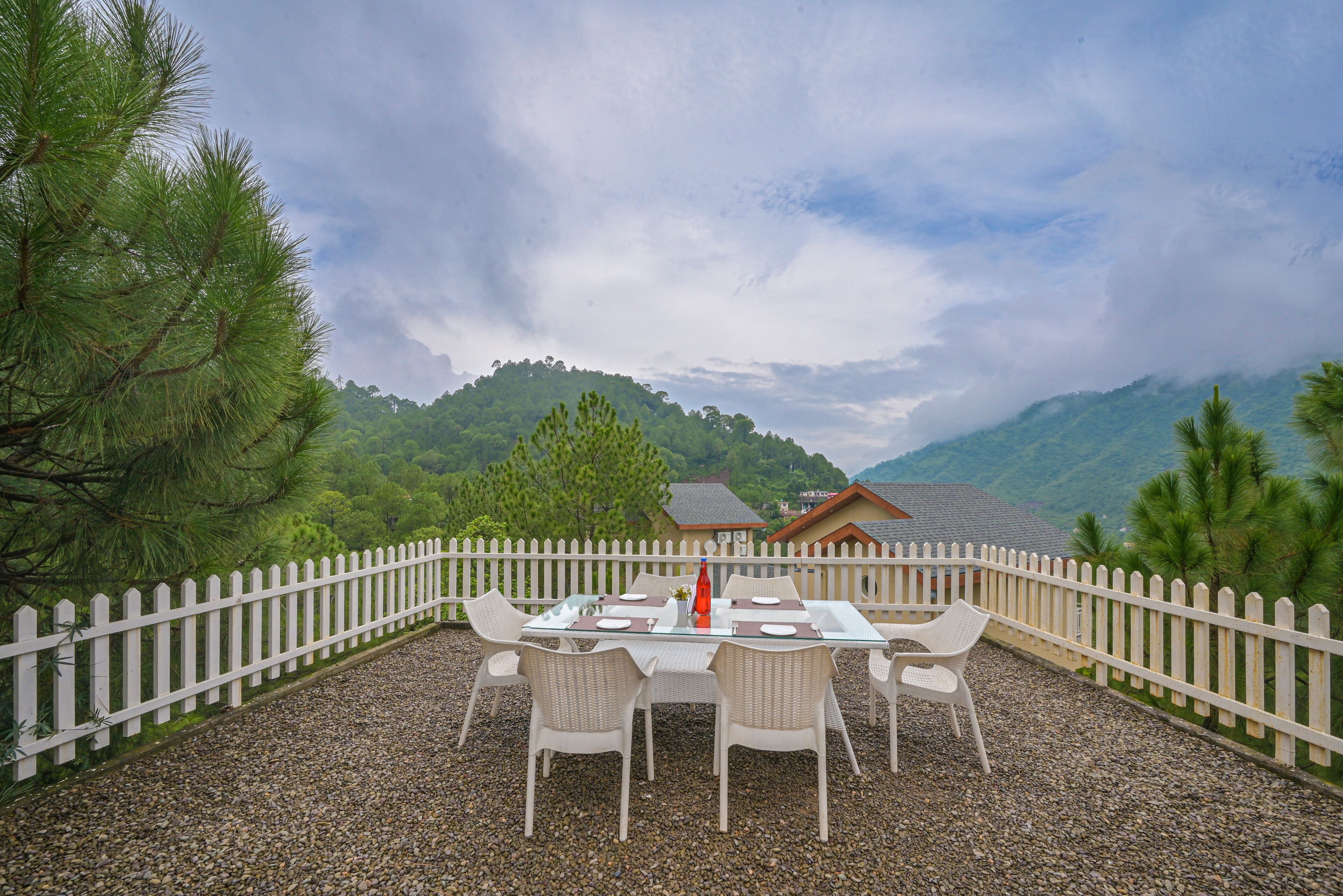 An outdoor dining area with a white picket fence, a table set for four, and a view of a mountain landscape.