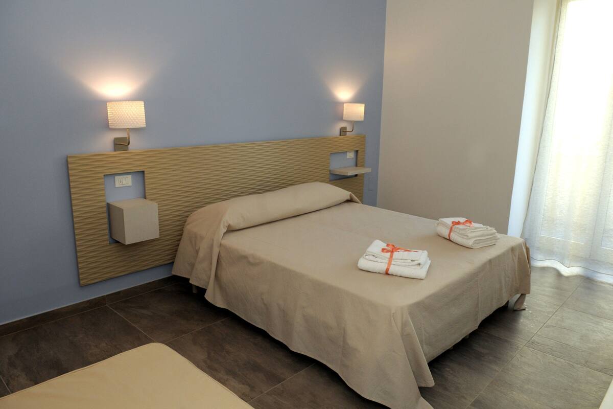 A neatly made bed with a beige bedspread and pillows, two bedside lamps, and a small shelf with a light fixture.