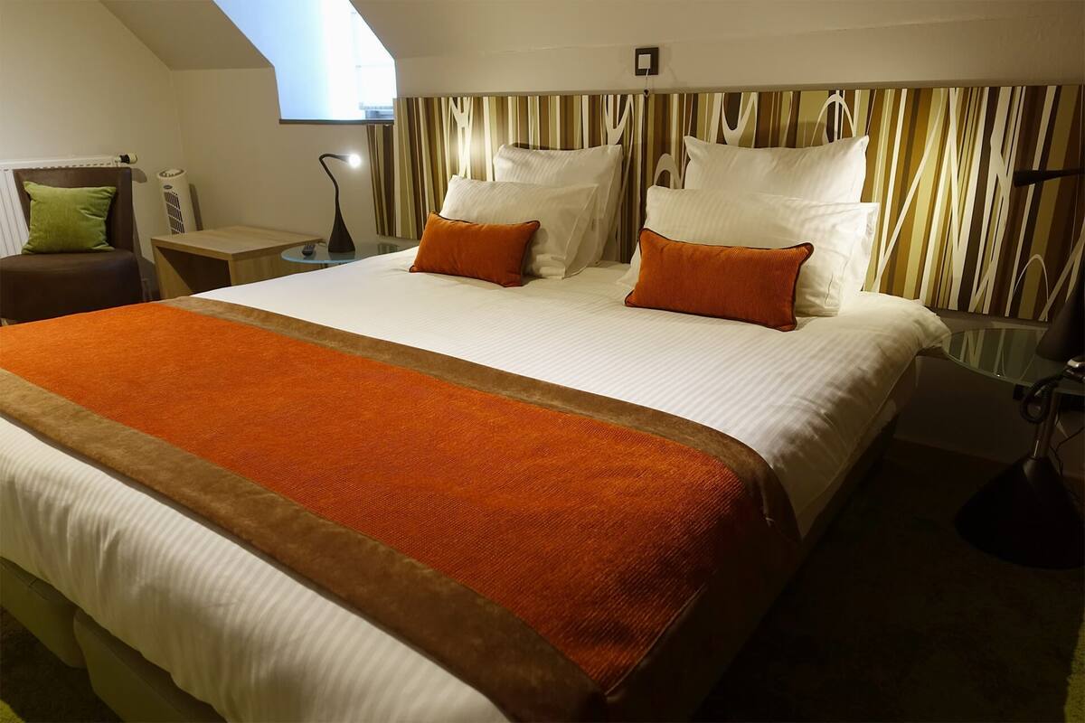 A hotel room with a large bed, a bedside table, a lamp, and a view of the sky through a skylight.