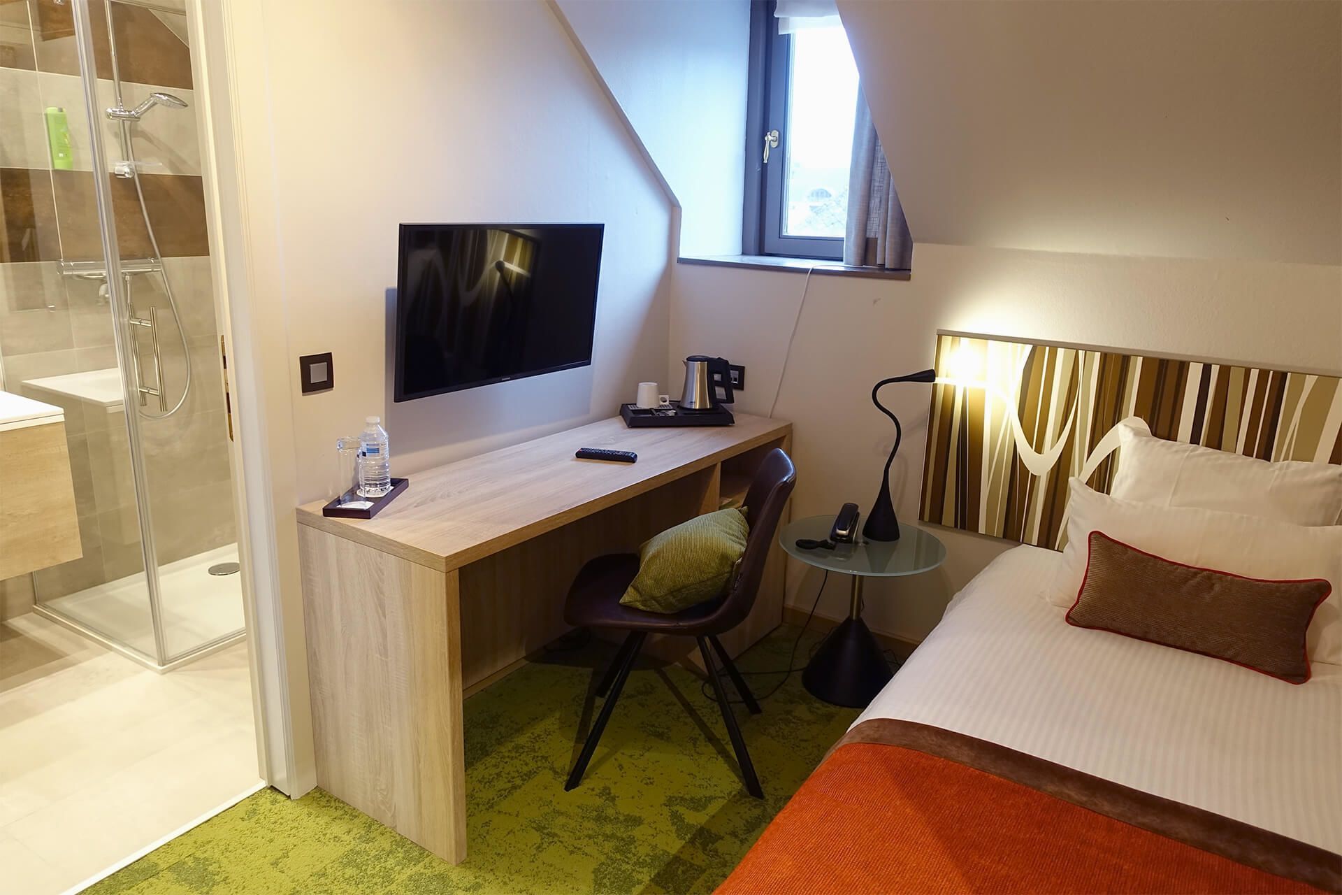 A hotel room with a shower, a desk with a TV, a bed, and a small table with a lamp.