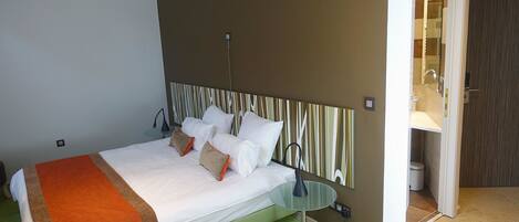 A modern hotel room with a bed, a glass table, and a bathroom visible through an open door.