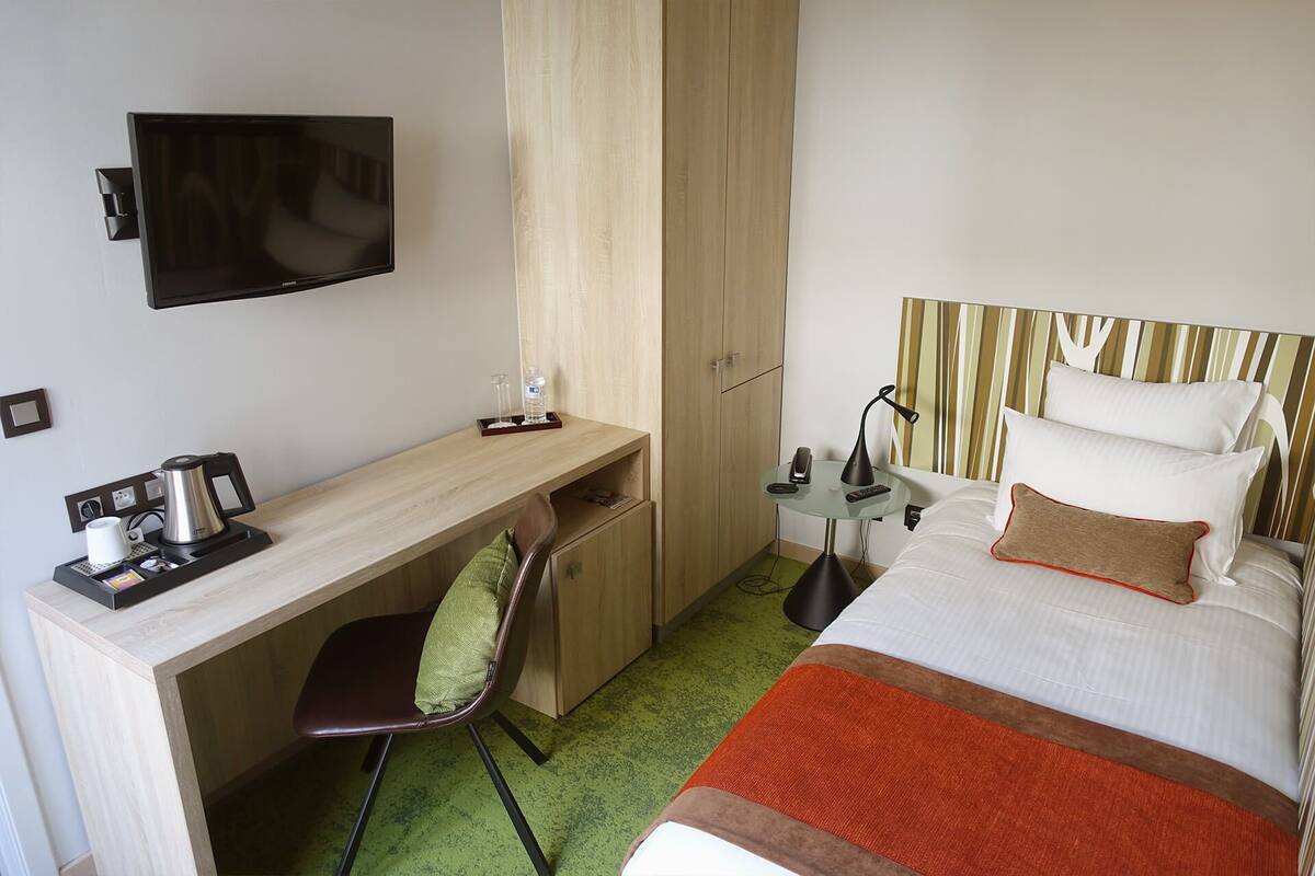 A hotel room with a bed, a desk with a chair, a TV, a bedside table with a lamp, and a wardrobe.