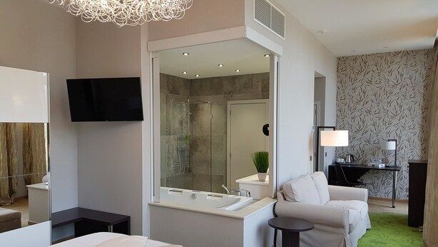 A modern hotel room with a large bed, a sitting area with a chair, a desk with a lamp, a shower area, and a TV mounted on the wall. - Hôtel du Parc (Ath)