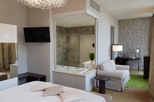 Habitación de hotel moderna con una cama grande, una zona de estar con una silla, un escritorio con una lámpara, un espacio de ducha y un televisor montado en la pared.