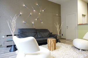 Una sala de estar moderna con un sofá de cuero negro, sillas blancas, una mesa de centro de vidrio y una pared decorativa con luces.