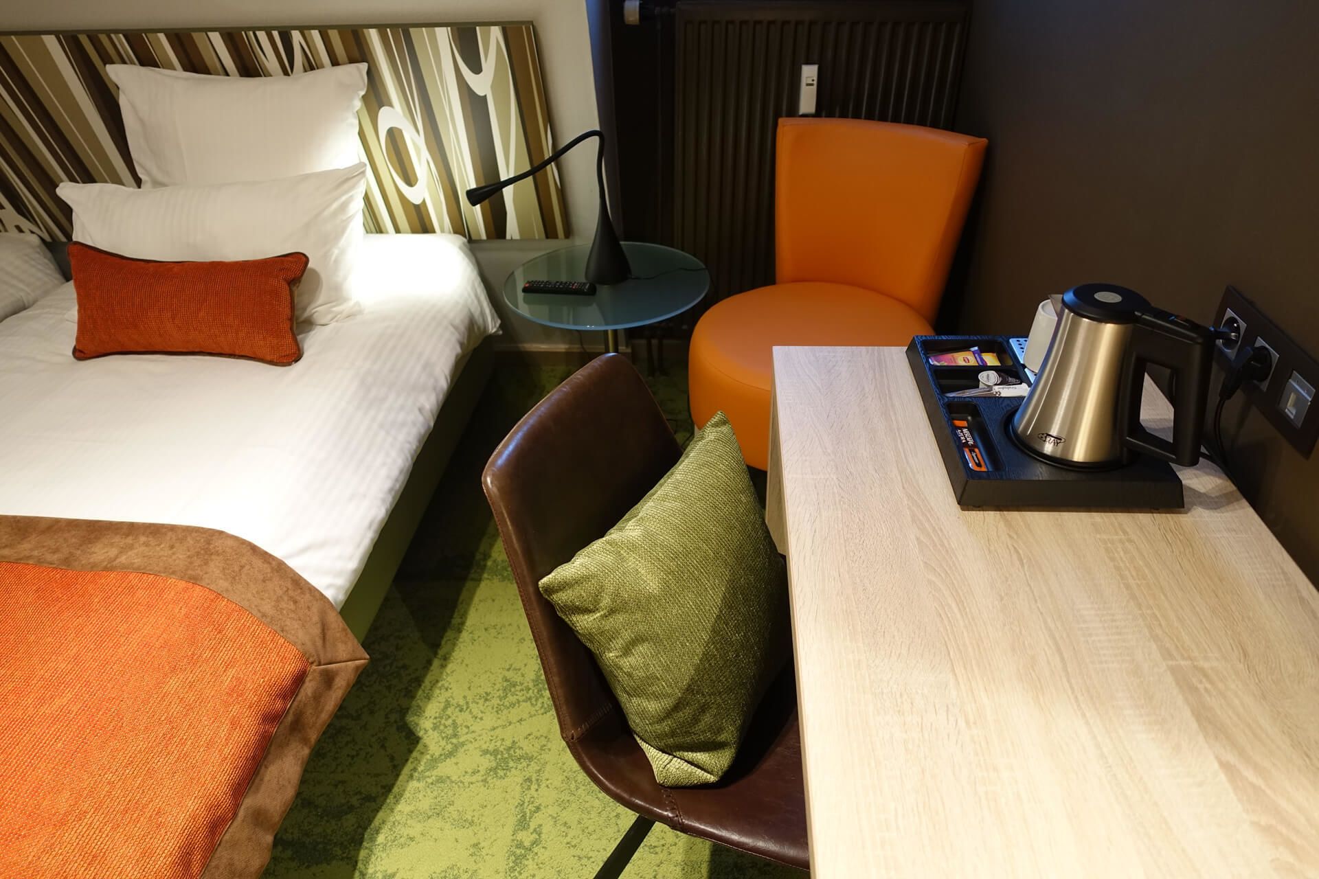 A hotel room with a bed, a desk with a kettle, a chair, and a small table.