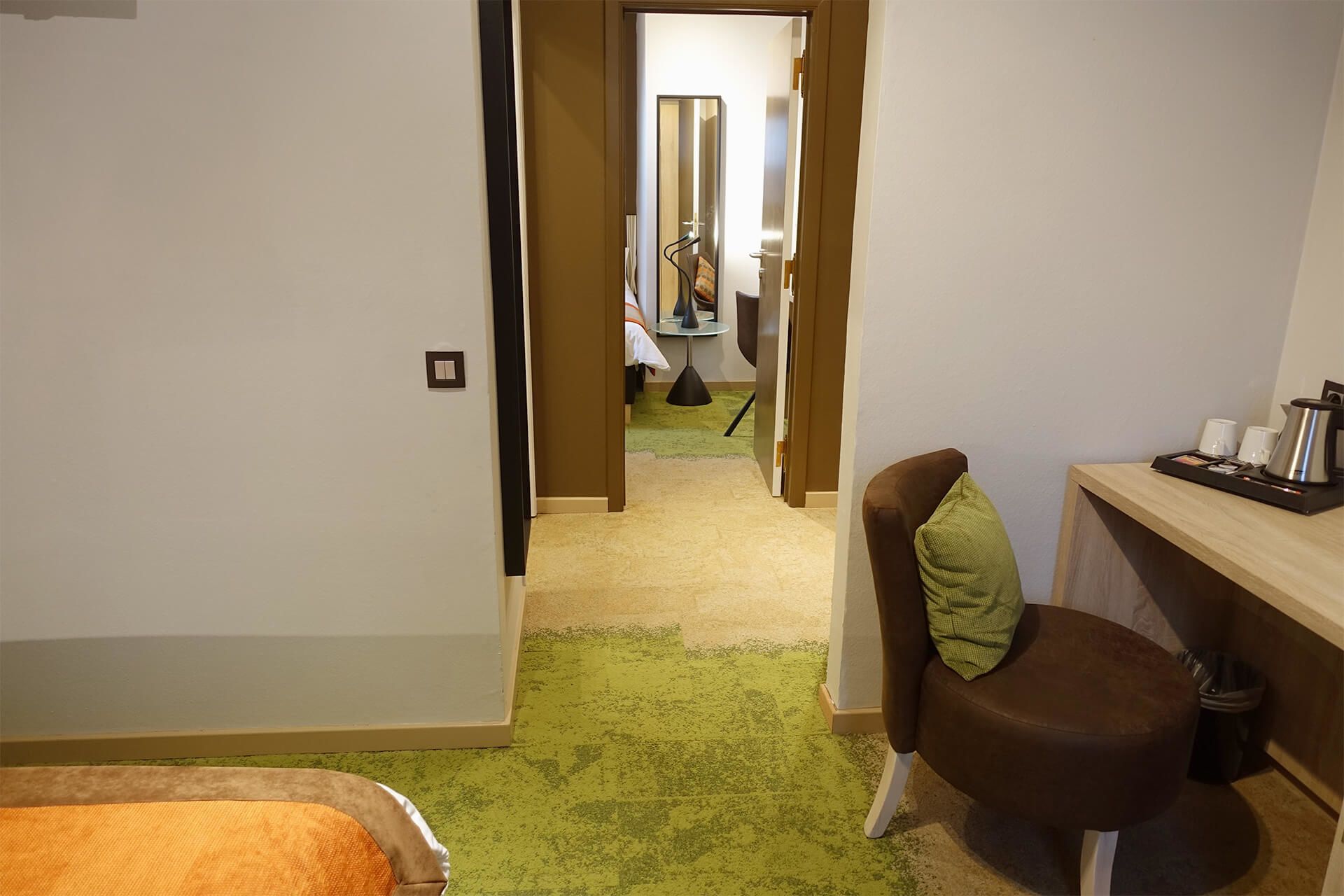 Un pasillo estrecho con una alfombra verde que conduce a una habitación con una cama, una silla y una mesita con una tetera.