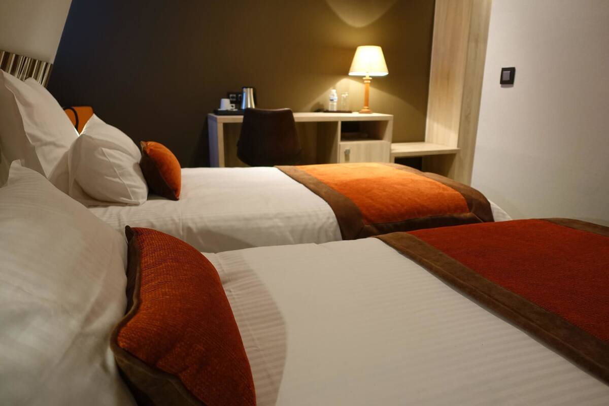 A hotel room with two beds, each with white linens and orange accents. There is a desk with a lamp, a chair, and a small table with a water bottle and a coffee maker.