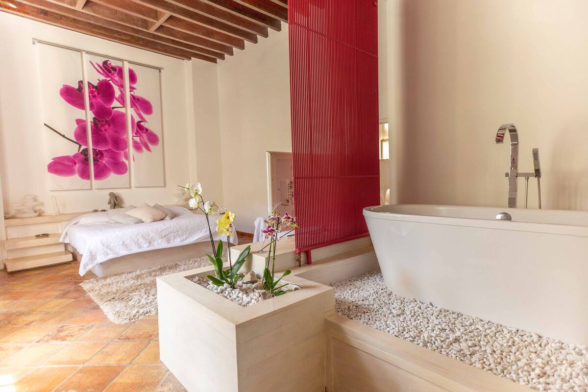 A modern bathroom with a large bathtub, a shower, and a toilet. There is a bed with a white bedspread and pillows, and a small table with plants.