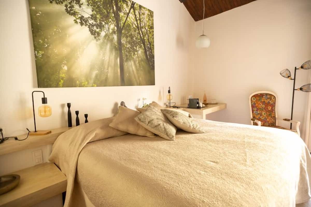 A bedroom with a large bed, a nightstand, a desk with a lamp, a chair, and a large forest-themed painting on the wall.