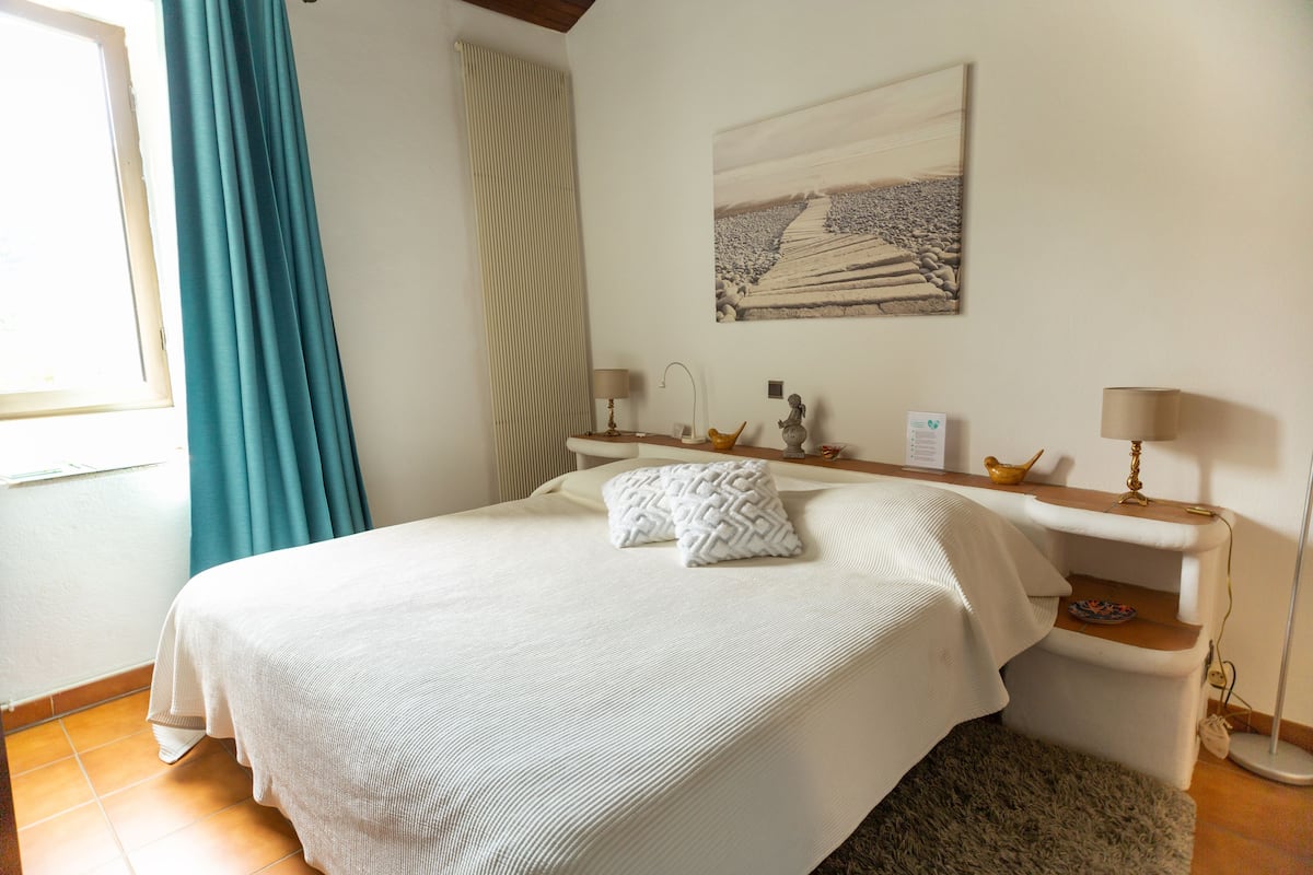 A neatly made bed with a patterned pillow, a bedside table with a lamp, a framed picture on the wall, and a window with curtains.