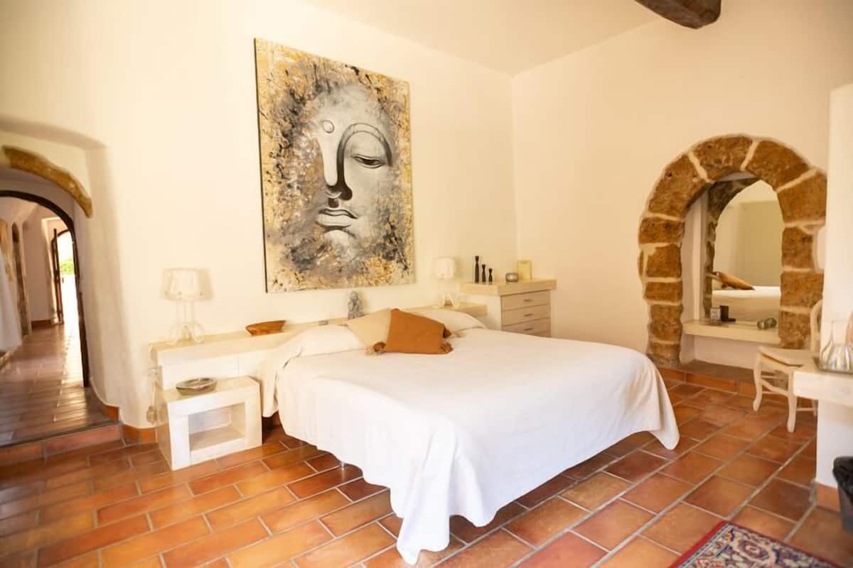 A bedroom with a large bed, a nightstand, a dresser, a mirror, and a stone archway.