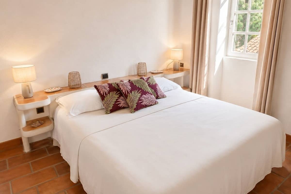 A neatly made bed with white bedding and decorative pillows, bedside tables with lamps, and a window with curtains.
