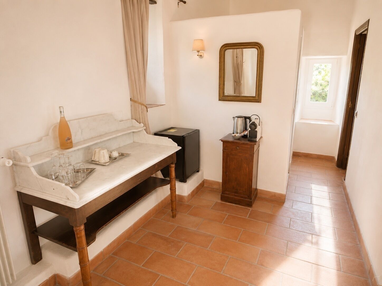 A room with a marble countertop, a bottle, glasses, a kettle, a coffee maker, a mirror, and a door leading to another room.