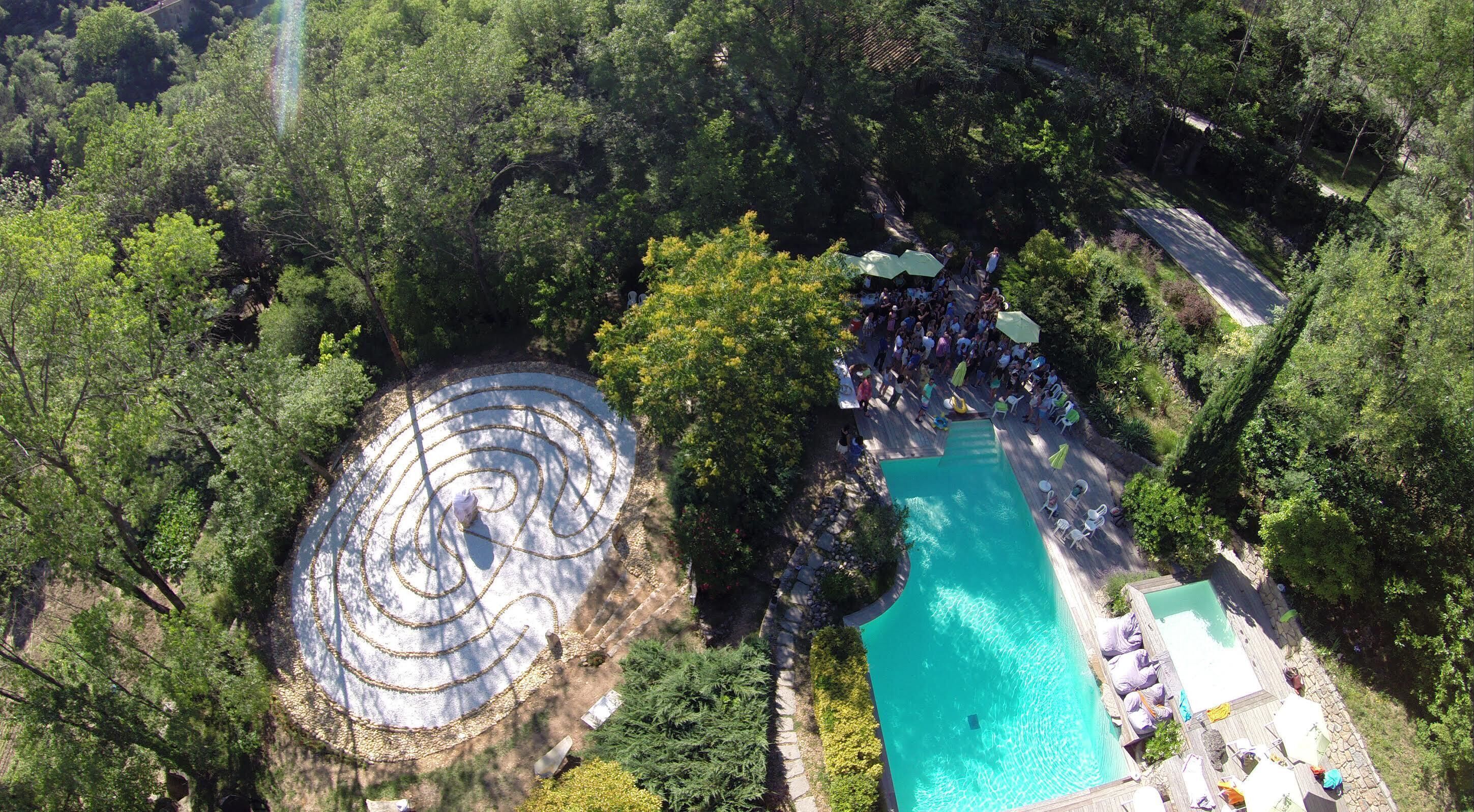 An aerial view of a resort with a swimming pool, a circular labyrinth garden, and lush greenery.