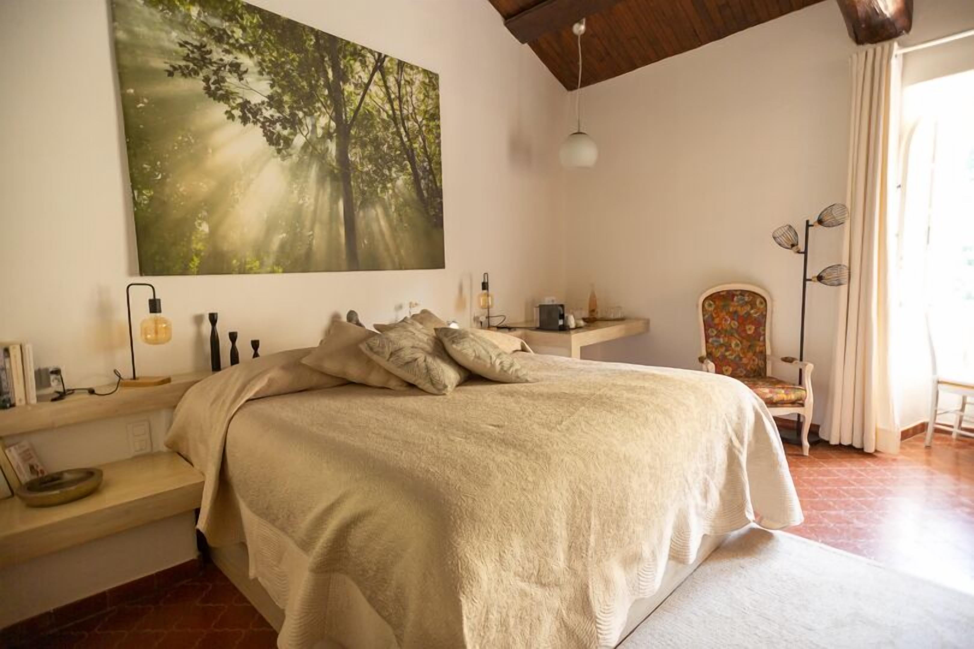 A bedroom with a large bed, a bedside table, a chair, and a painting of a forest.