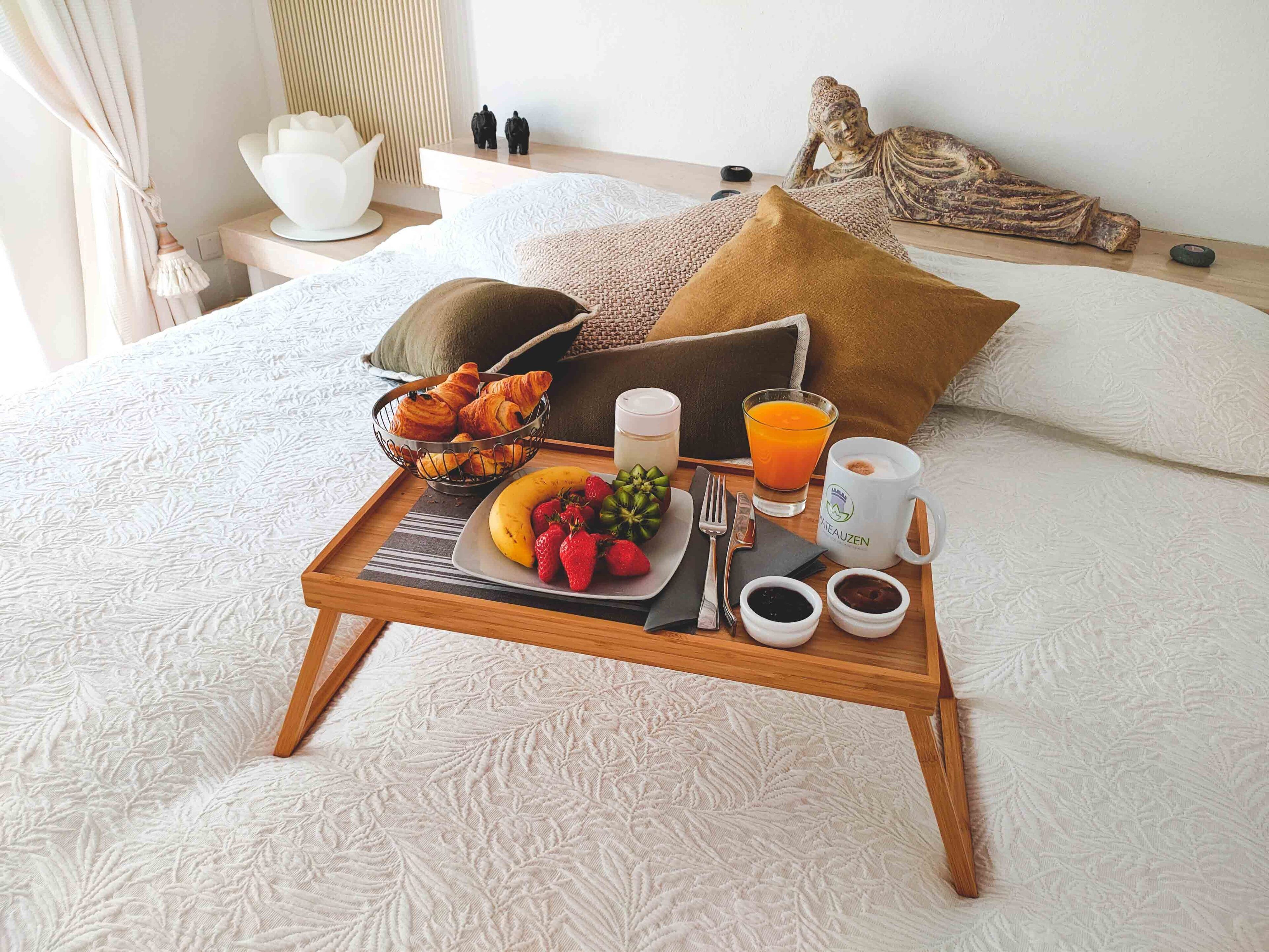 Un plateau de petit-déjeuner, joliment disposé, est posé sur un lit recouvert d’une couverture blanche à texture particulière. On y trouve un bol de croissants, une assiette de fruits, un verre de jus d’orange et deux tasses.