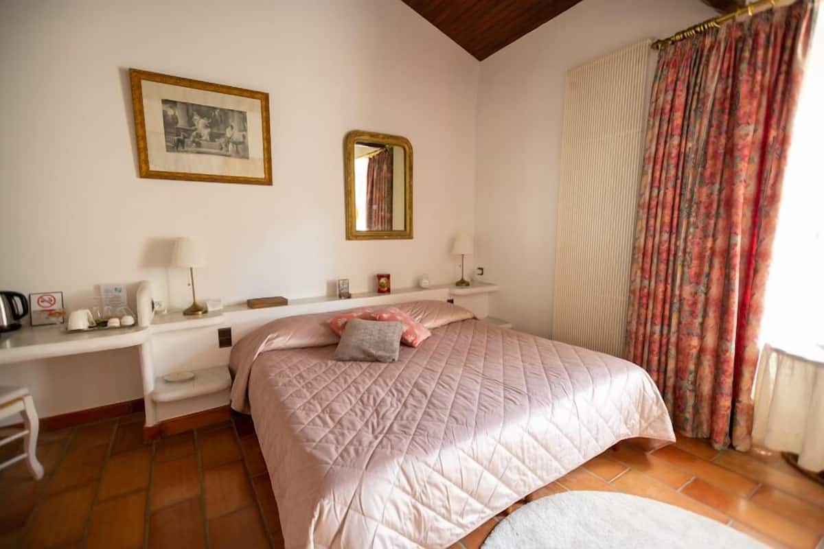 A bedroom with a bed, a bedside table, a mirror, a picture frame, and a window with curtains.