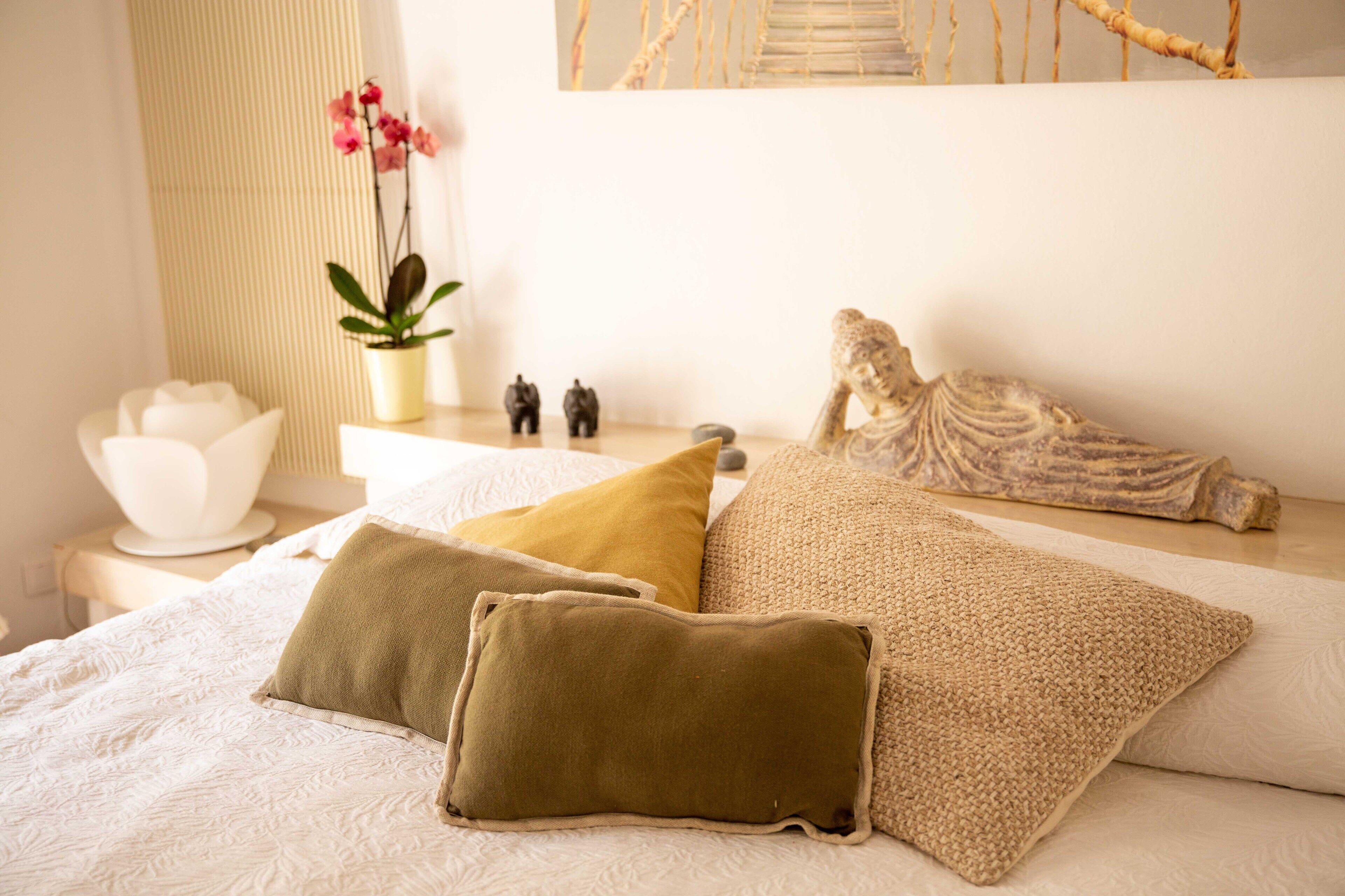A neatly made bed with three pillows, a small white table with a bowl and figurines, and a potted plant with pink flowers.