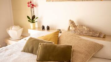 A neatly made bed with three pillows, a small white table with a bowl and figurines, and a potted plant with pink flowers.