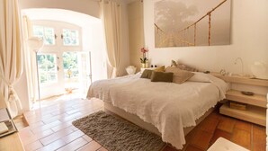 Un dormitorio con una cama grande, piso de madera, ventana con cortinas, mesita de noche con lámpara y una hamaca fija a la pared.