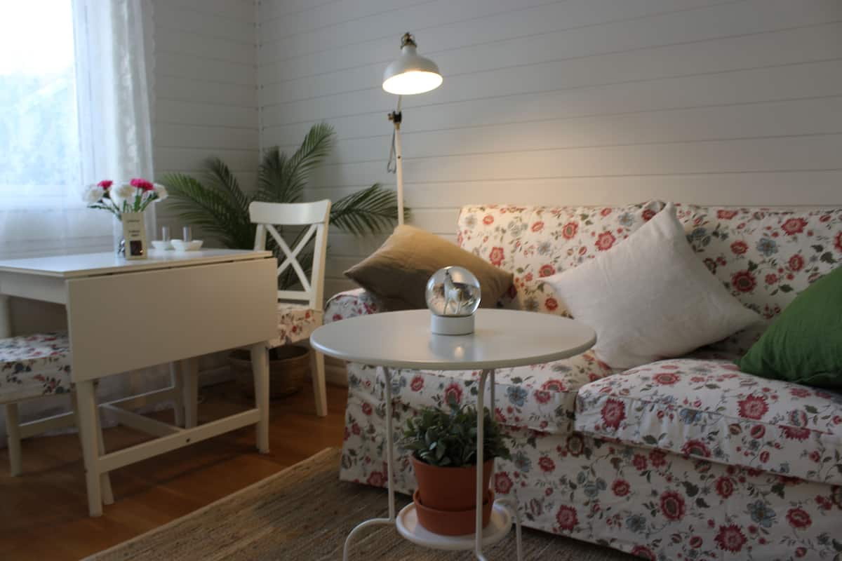 A cozy living room with a floral sofa, a small round table, a white desk, and a floor lamp.