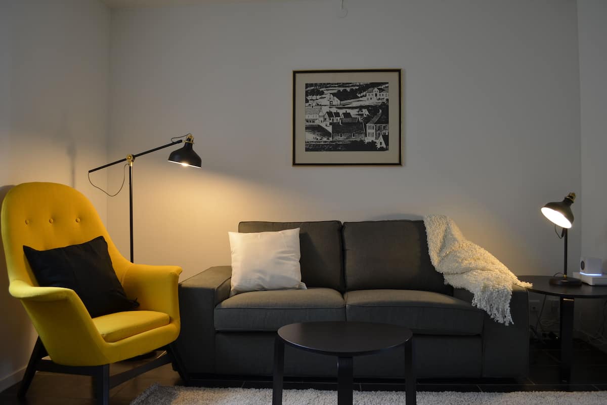 A living room with a yellow armchair, a grey sofa, a black floor lamp, and a framed black and white photograph on the wall.
