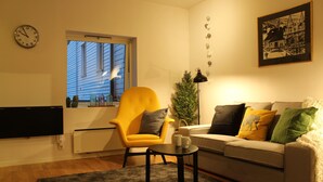 Una sala de estar con un sillón amarillo, un sofá gris, una mesa de centro negra y una ventana con persianas.