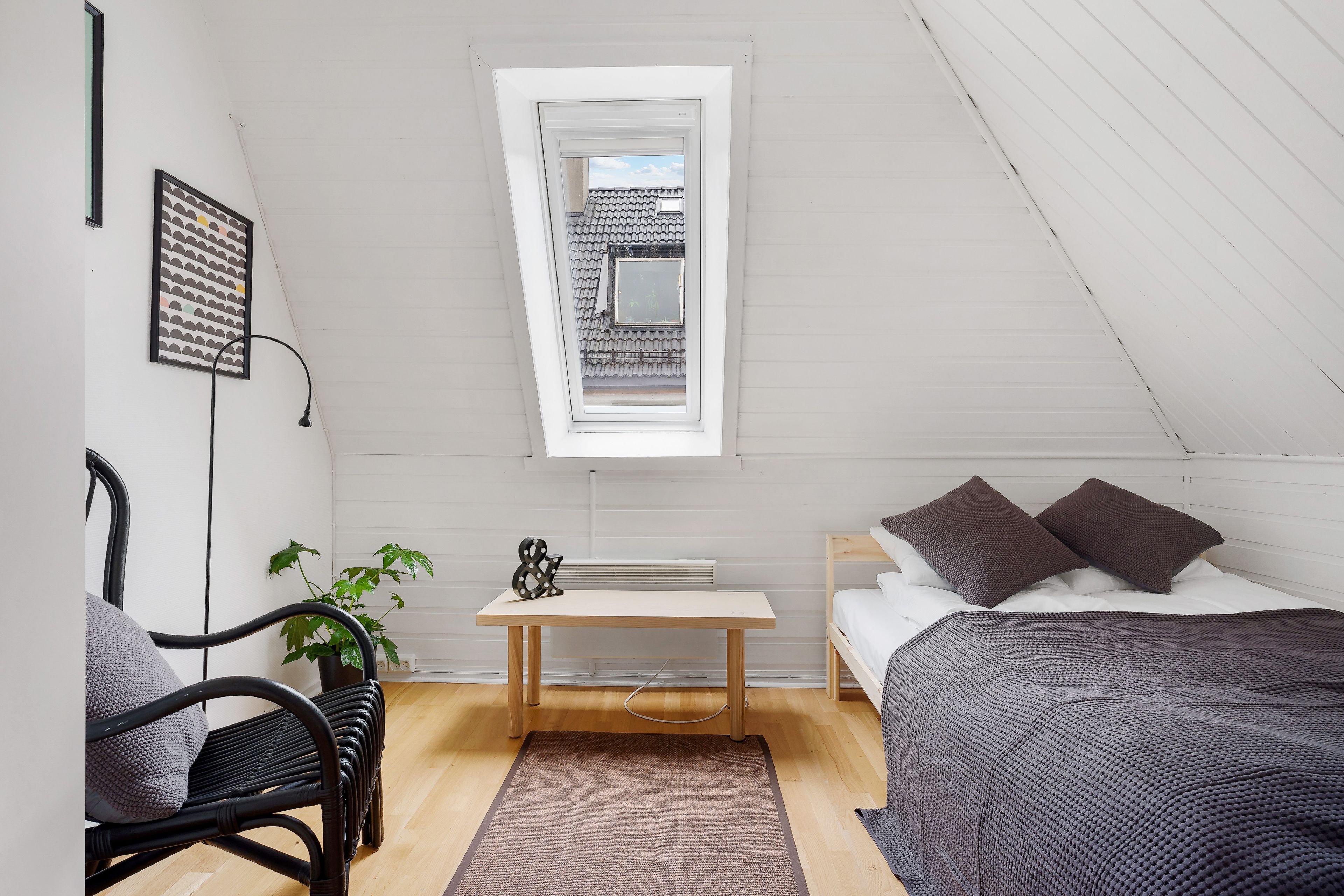 A bedroom with a sloped ceiling, a skylight, a bed with dark bedding, a bedside table with a lamp, a chair, and a potted plant.
