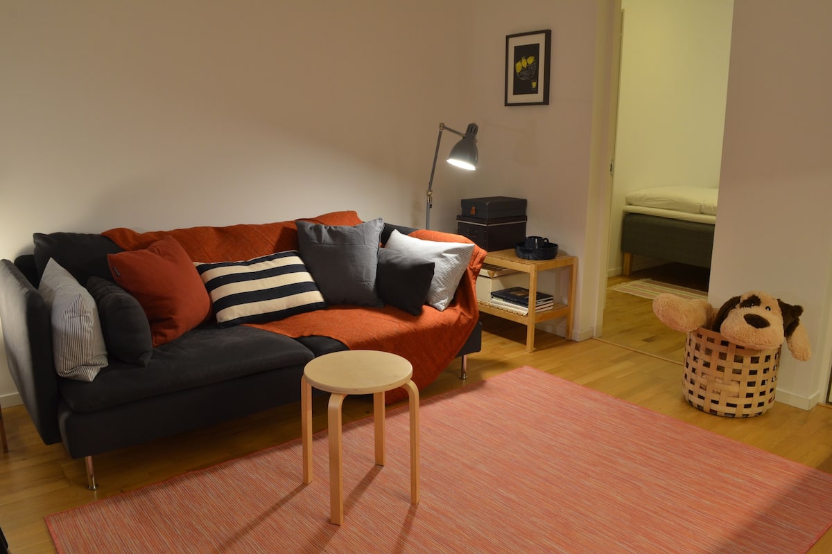 A cozy living room with a sofa, a small wooden table, a lamp, a plush toy, and a bed in the background.