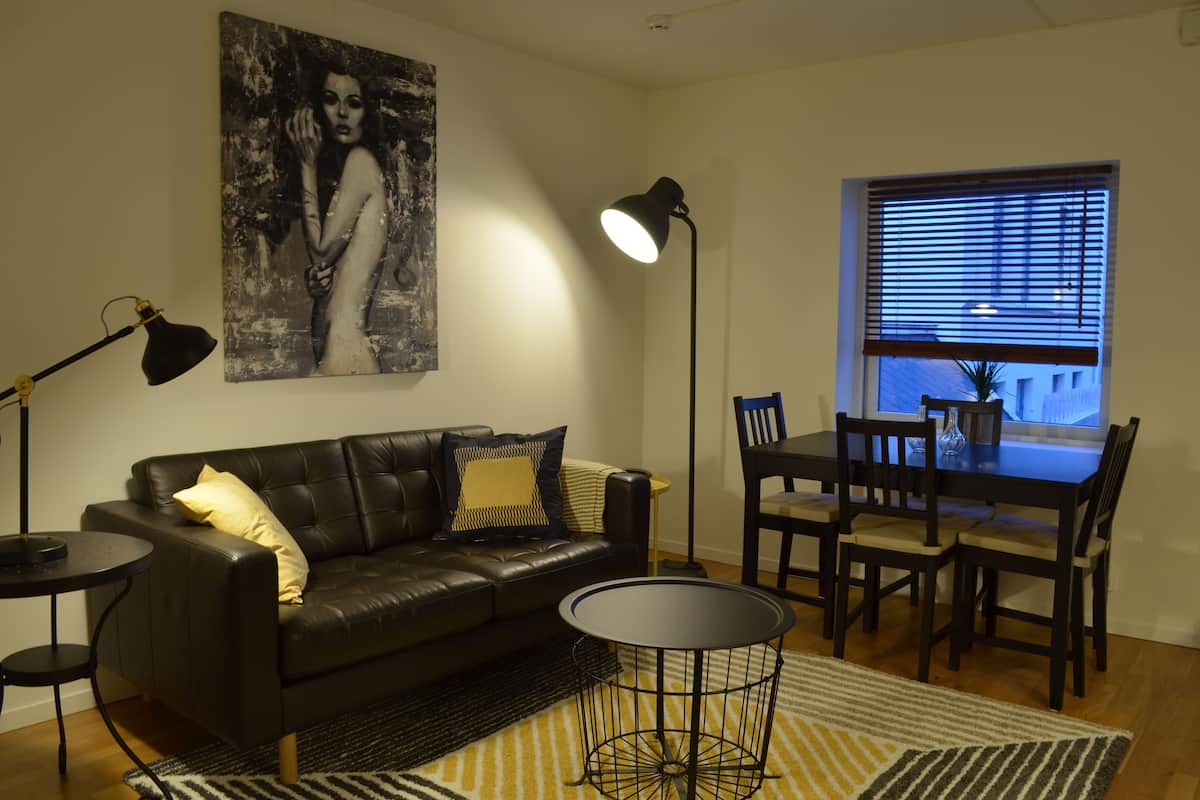 A modern living room with a black leather sofa, a dining table with chairs, a black floor lamp, and a large abstract painting on the wall.