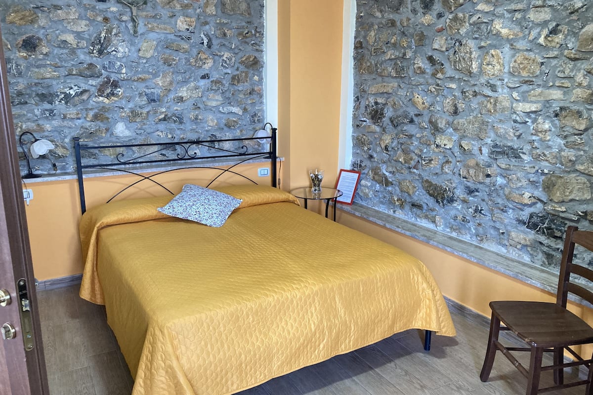 A bed with a yellow bedspread, a stone wall, a small table with a glass, and a wooden chair.