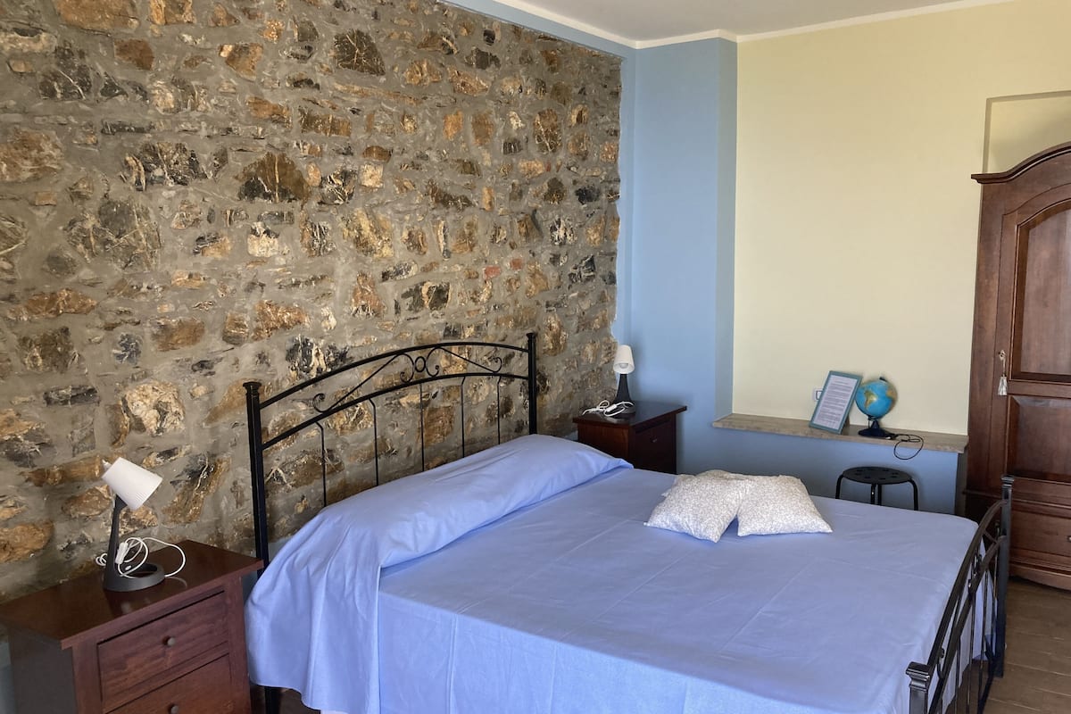 A bedroom with a stone wall, a bed with a blue bedspread, a nightstand with a lamp, a wooden dresser, and a desk with a globe and a framed document.