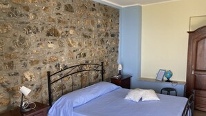 A bedroom with a stone wall, a bed with a blue bedspread, a nightstand with a lamp, a wooden dresser, and a desk with a globe and a framed document. - Relais da Clorinda (Ascea)