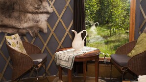 Une pièce avec un fauteuil en osier, une table sur laquelle se trouve une carafe, et une vue sur la verdure à l’extérieur.