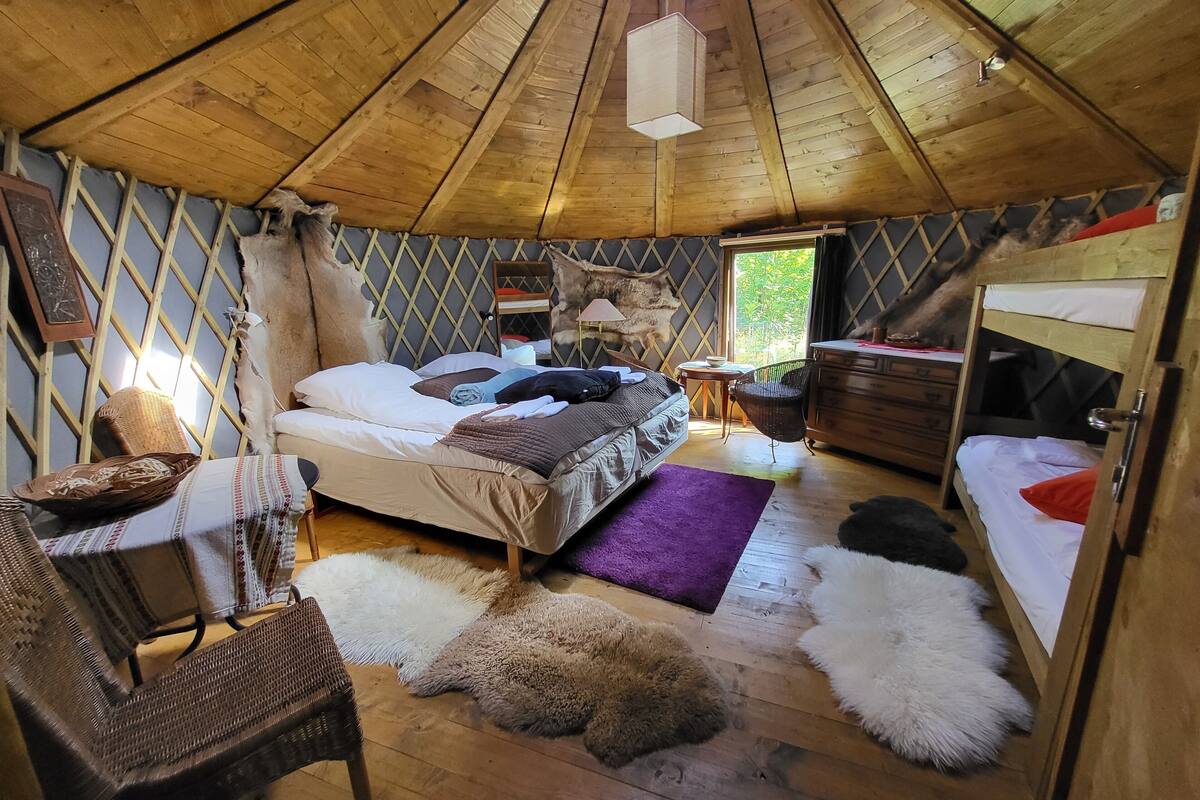 A cozy, rustic room with a high wooden ceiling, a bed with a patterned bedspread, a wicker chair, a small wooden table, and a window with a view of greenery.