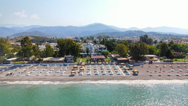 Vista aerea di una spiaggia con file di lettini e ombrelloni, un grande edificio sullo sfondo e un paesaggio montuoso.