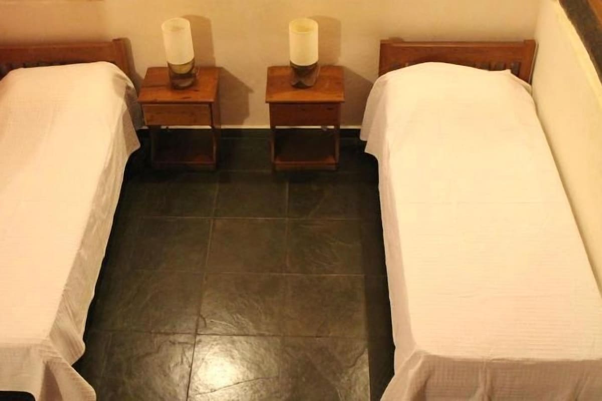 Una habitación pequeña con dos camas individuales, cada una con cabecero de madera y una mesita de noche con lámpara. El suelo está cubierto de baldosas.