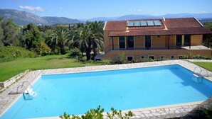 Une maison à deux étages avec une piscine, entourée de verdure et avec des montagnes en toile de fond.