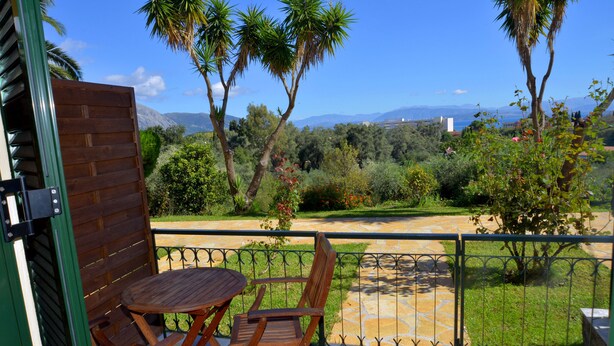 Un balcon avec une table et des chaises en bois, un chemin de pierre et une vue sur un paysage verdoyant, avec des arbres et des montagnes à l’horizon.