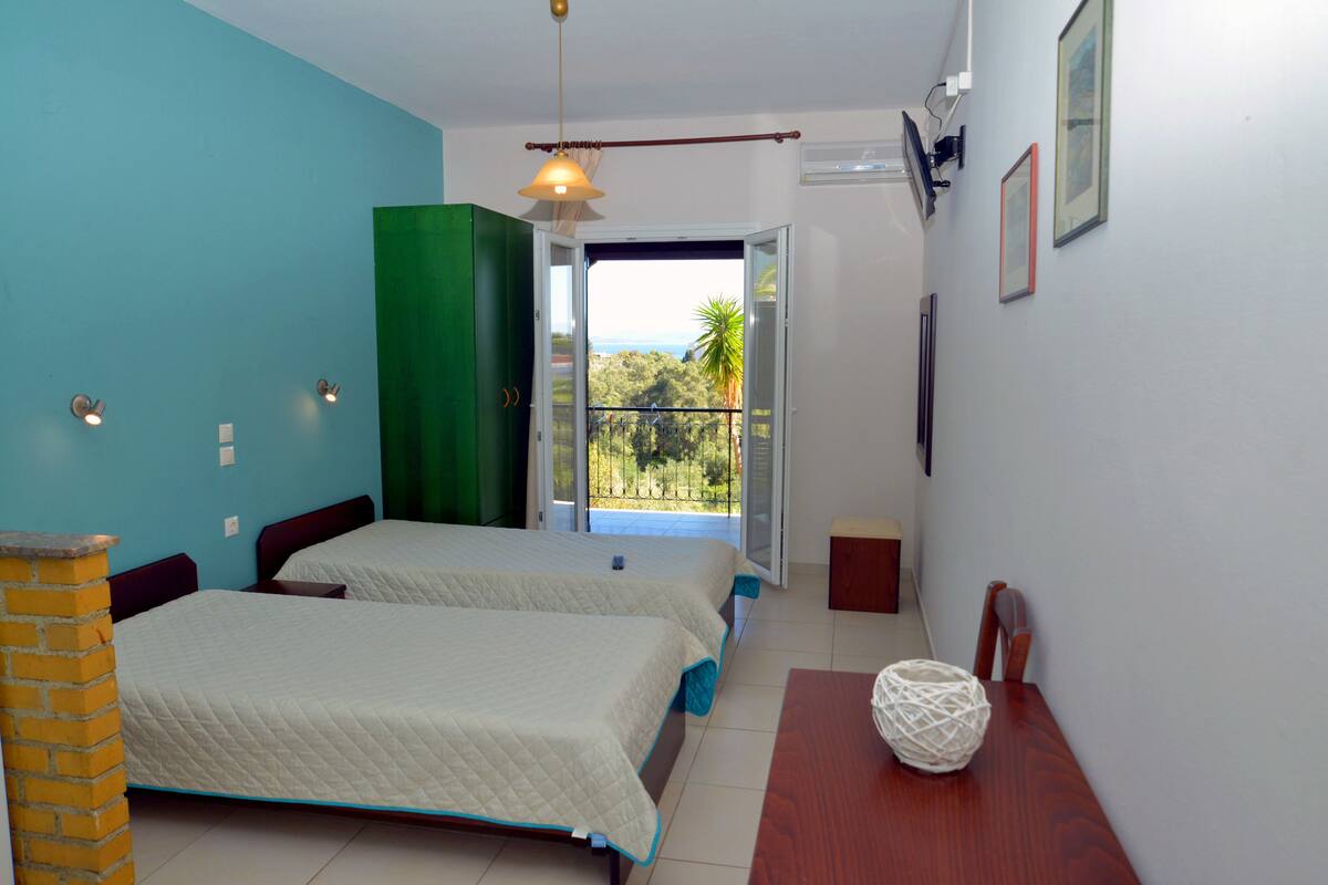 A bedroom with two beds, a green wardrobe, a wooden table, and a view of the outdoors.