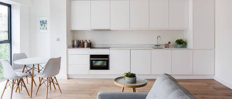 A modern kitchen with white cabinets, a built-in oven, and a sink. There is a dining area with a white table and chairs, and a gray sofa in the living room.