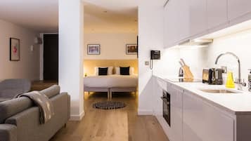 Camera d'albergo compatta con angolo cottura, un letto con biancheria in bianco e nero, un divano grigio e un piccolo tappeto.
