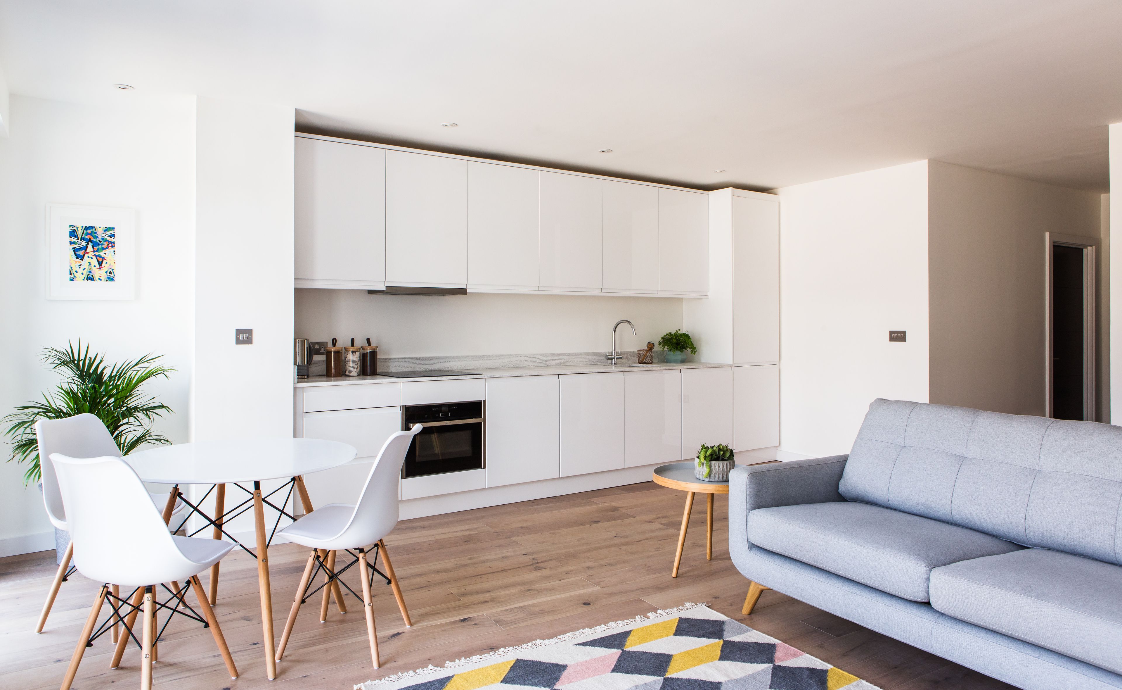 A modern kitchen with white cabinets, a marble countertop, and a built-in oven. There is a dining area with a white table and chairs, and a grey sofa in the living room.