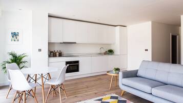 A modern kitchen with white cabinets, a marble countertop, and a built-in oven. There is a dining area with a white table and chairs, and a grey sofa in the living room.