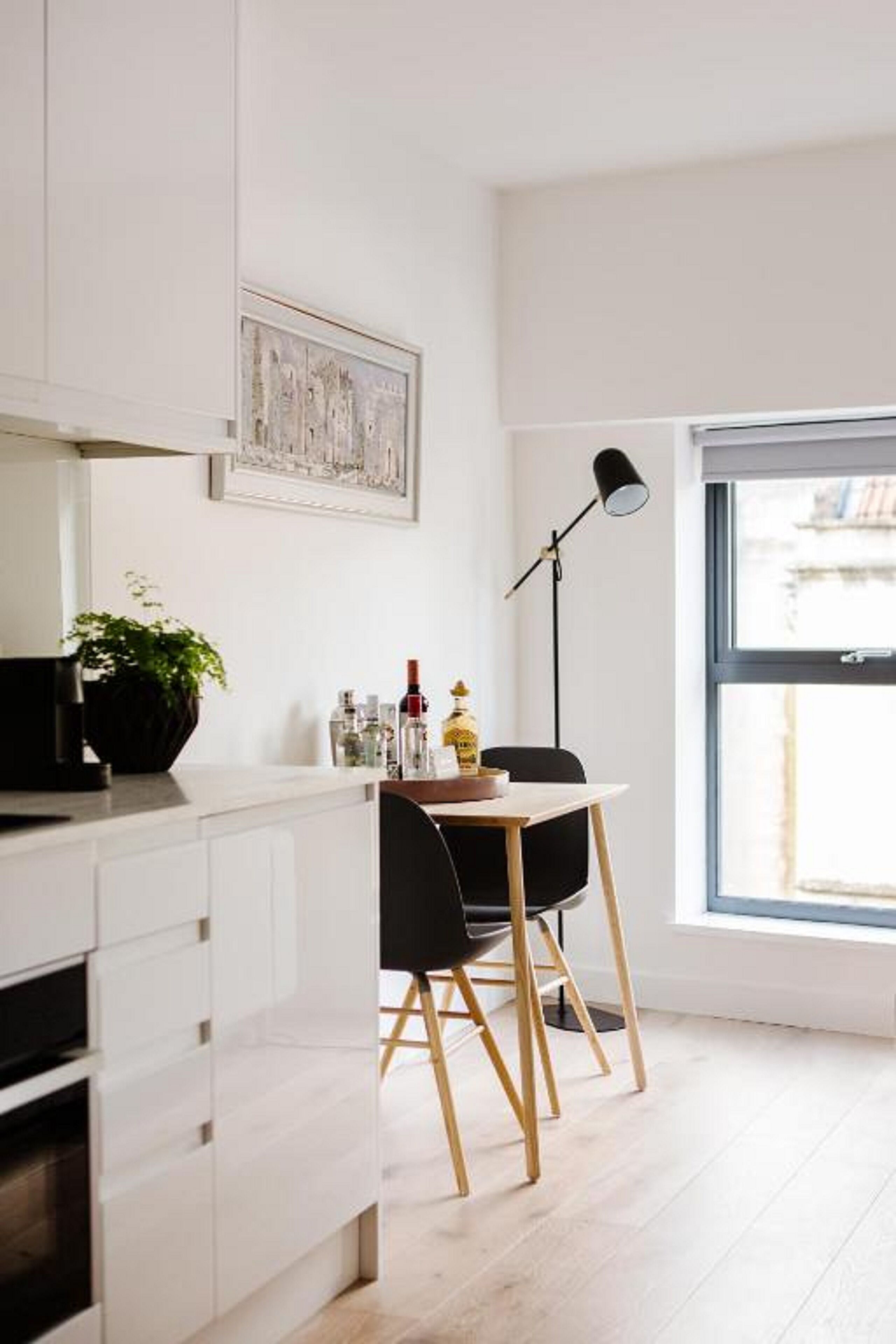 Eine moderne Küche mit weißen Schränken, einer schwarzen Arbeitsplatte, einem Holzstuhl, einer Stehlampe und einem Fenster mit Blick nach draußen.